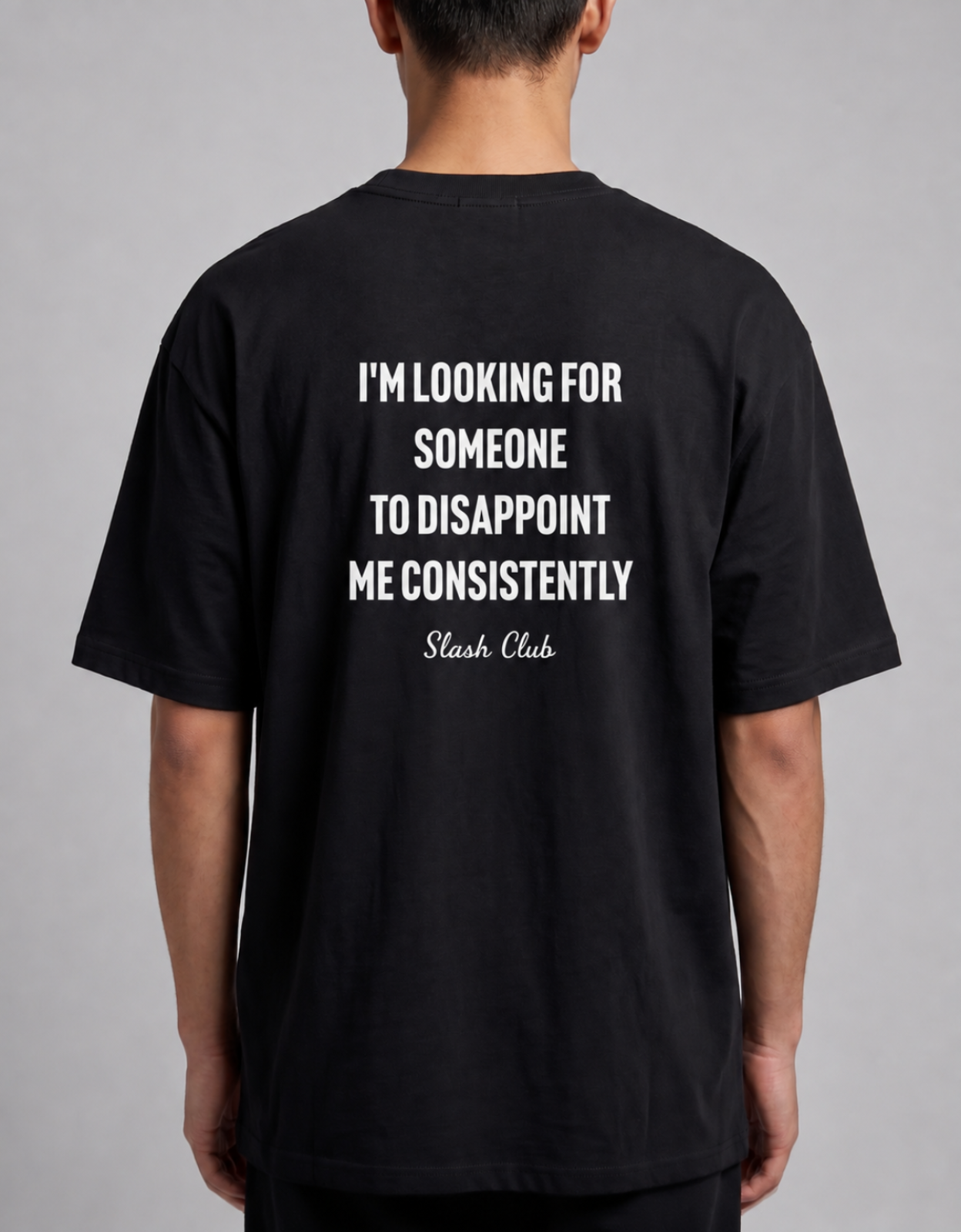 A man viewed from the back wearing a black t-shirt with a bold white text graphic on the back that reads 'I'M LOOKING FOR SOMEONE TO DISAPPOINT ME CONSISTENTLY Slash Club'.