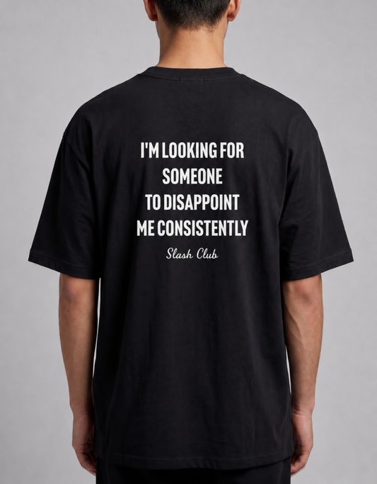 A man viewed from the back wearing a black t-shirt with a bold white text graphic on the back that reads 'I'M LOOKING FOR SOMEONE TO DISAPPOINT ME CONSISTENTLY Slash Club'.